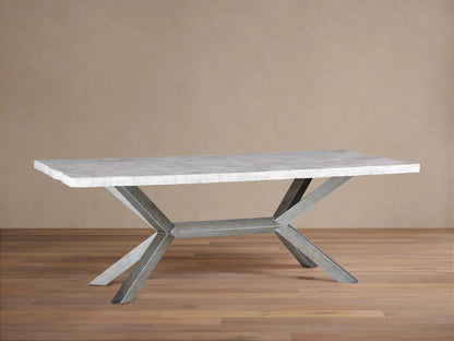 Petra Dining Table with a polished, light stone top and iron base in an X-shape, featuring a dark grey patina, placed on wooden flooring.