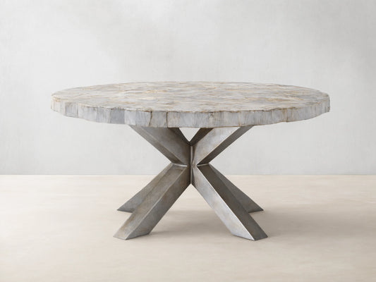 Round dining table with petrified wood top and textured surface, supported by an X-shaped iron base with dark grey patina, on wood floor.