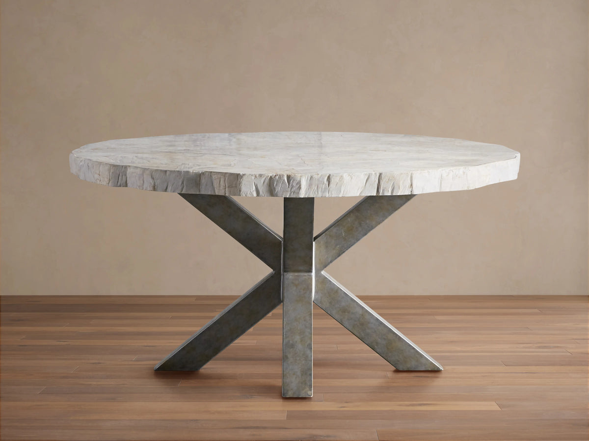 Round dining table with petrified wood top and textured surface, supported by an X-shaped iron base with dark grey patina, on wood floor.