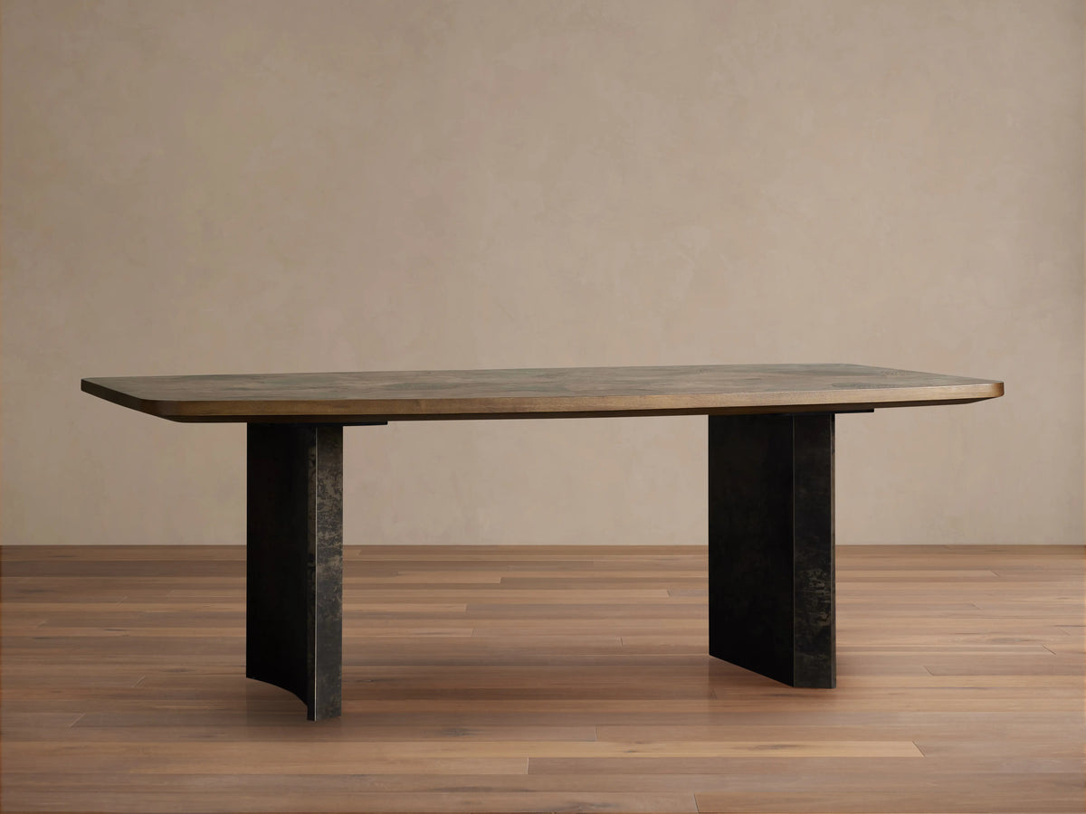 Polanco Dining Table with a mottled dark brown wooden top and two weathered dark metal, curved pedestal legs on a wooden floor.