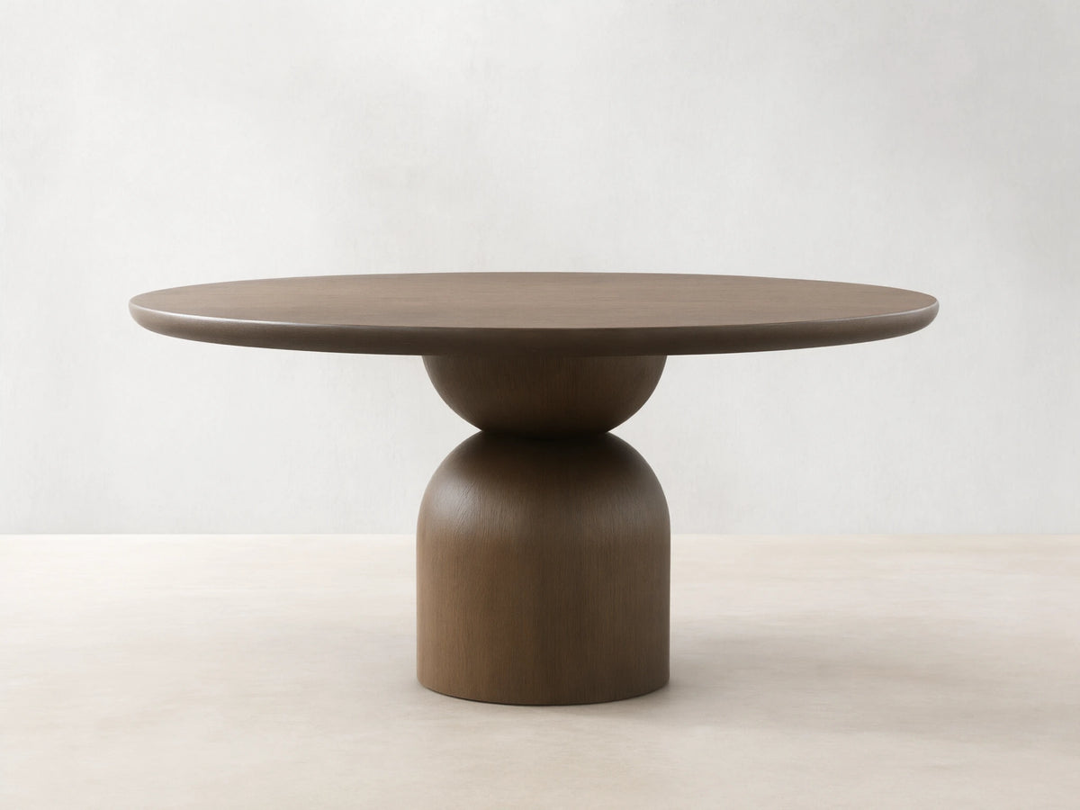 Dark wood Perth Round Dining Table with a thick circular top and a modern, sculptural pedestal base on a wood floor.
