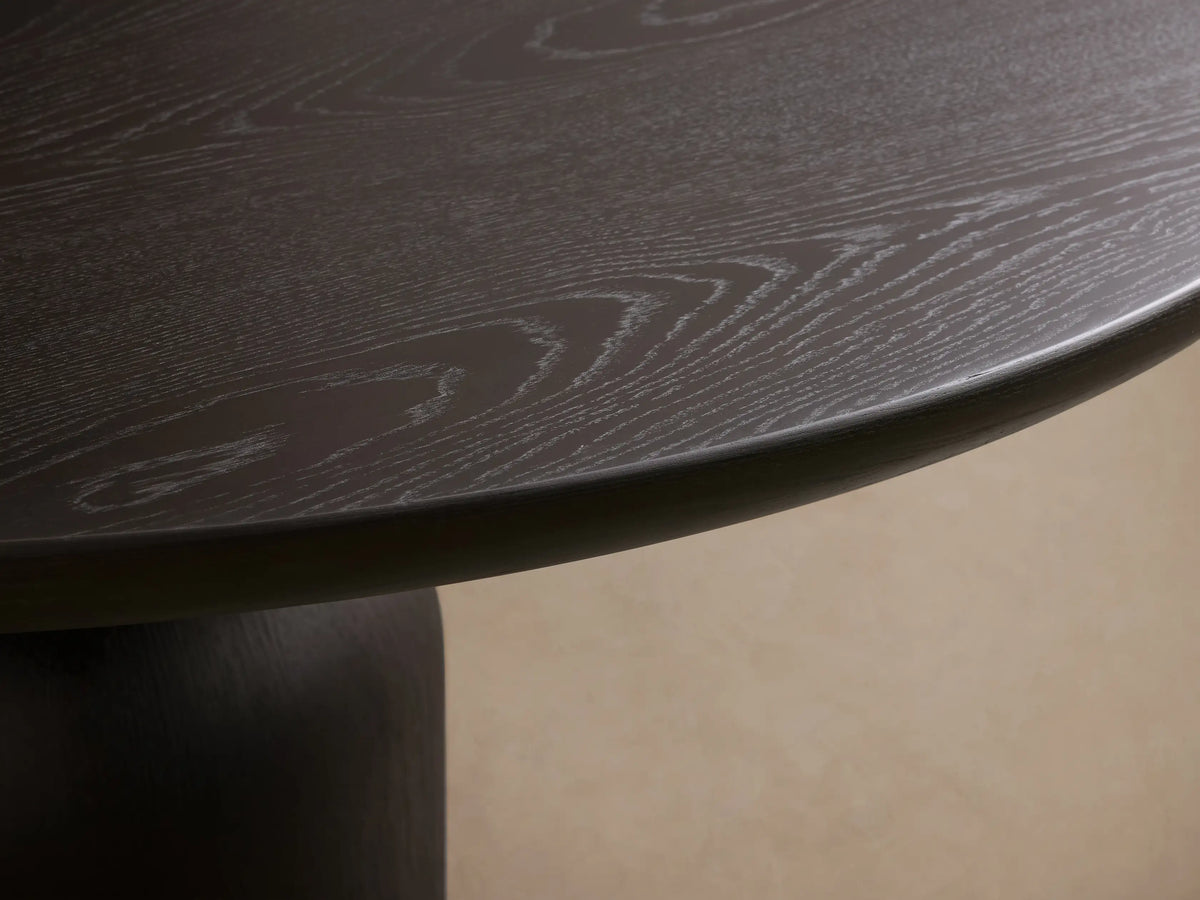 Dark brown round dining table with a sculptural hourglass pedestal base, on a wood floor in front of a beige wall.