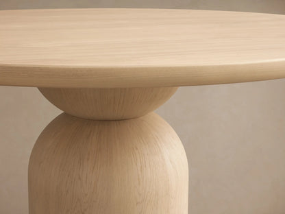 Perth Round Dining Table: Light oak wood, thick circular top, and a distinctive sculptural rounded base, on a wooden floor.