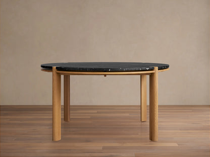 Pyet Round Dining Table with a black marble top and four round, natural red oak legs on a wooden floor, set against a beige wall.