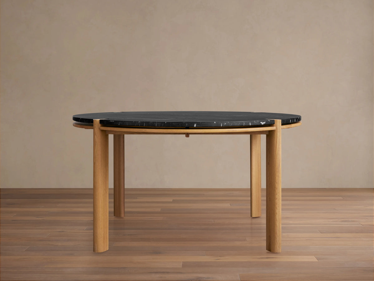 Pyet Round Dining Table with black marble top and natural red oak wood legs. Table is set on a polished wooden floor against a neutral wall.