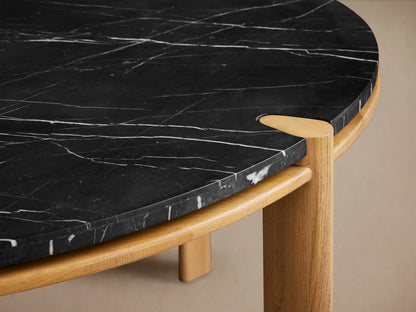 Pyet Round Dining Table with a black Carrara marble top and solid red oak base on a wooden floor background.