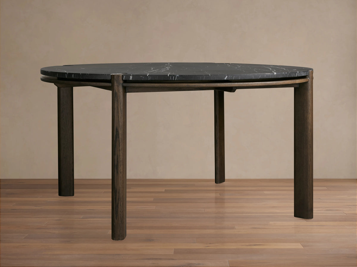 Round dining table with dark marble top, white veining, and brown wood accents visible at joints against a beige background.