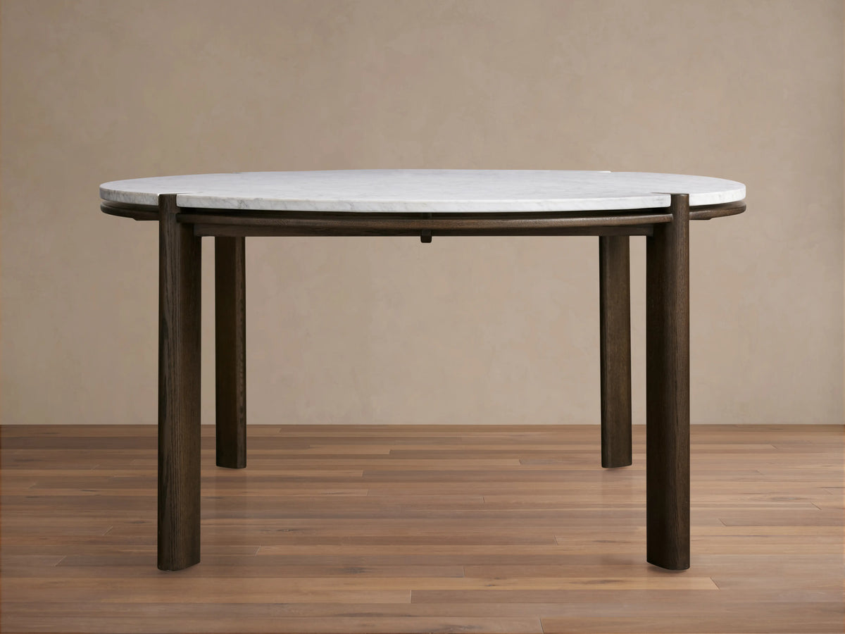 Pyet Round Dining Table with a white Carrara marble top and robust solid red oak legs, set on a wooden floor against a beige wall.