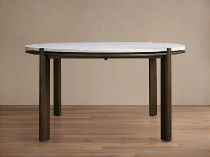 Pyet Round Dining Table with a white Carrara marble top and robust solid red oak legs, set on a wooden floor against a beige wall.