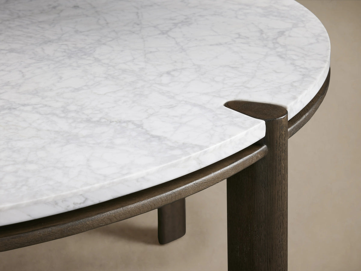 Pyet Round Dining Table with a white Carrara marble top and solid red oak wood legs, set in a room with a wooden floor.