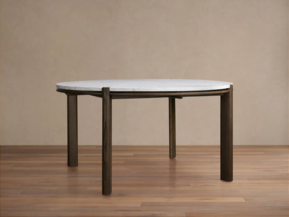 Pyet Round Dining Table with white marble top and dark red oak wooden legs, set on a wooden floor against a plain neutral wall.