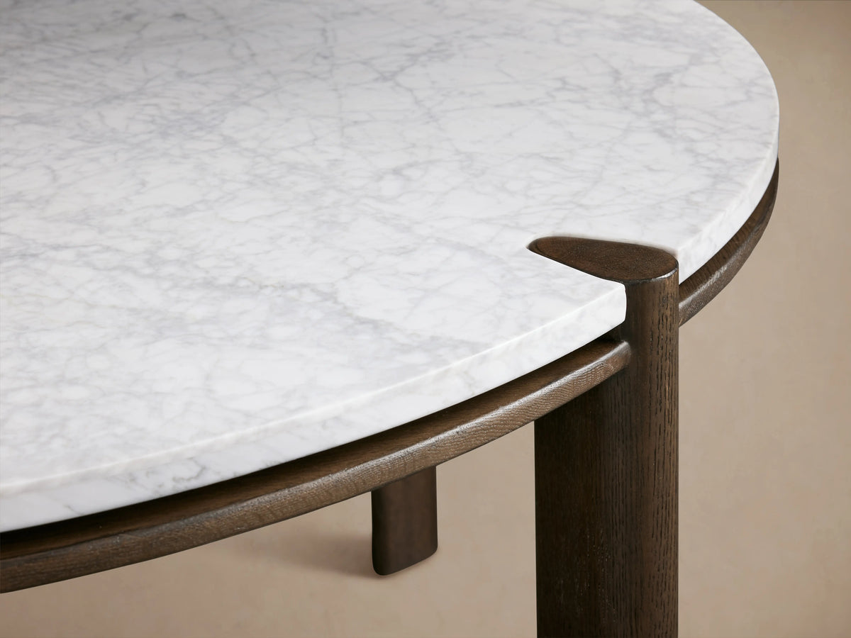 Pyet Round Dining Table with a white Carrara marble top and solid red oak wood legs, set on a wooden floor in a neutral-toned room.