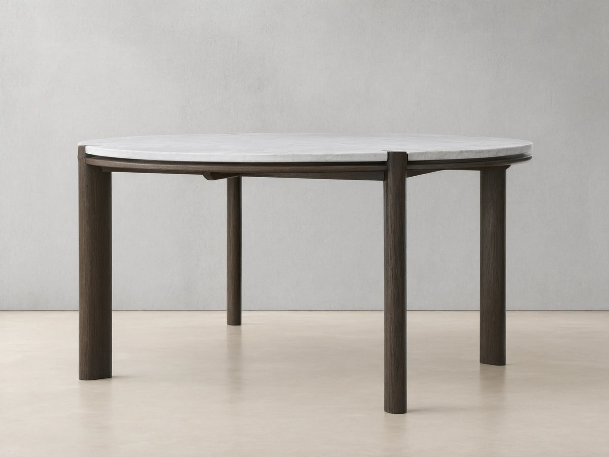 Pyet Round Dining Table with a white Carrara marble top and robust solid red oak legs, set on a wooden floor against a beige wall.