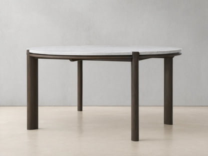 Pyet Round Dining Table with a white Carrara marble top and robust solid red oak legs, set on a wooden floor against a beige wall.