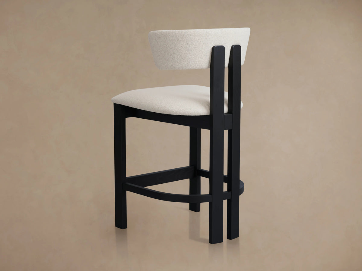 Rodin Armless Stool with cream-colored cushioned seat and backrest on a black ash wood frame, shown on a wooden floor.