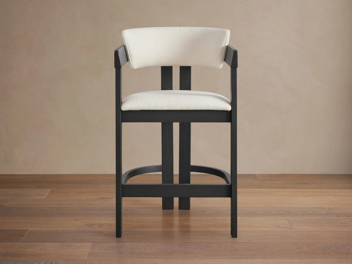 Rodin Counter Stool with cream upholstered seat and back, black ash wood frame, placed on a wooden floor against a beige wall.