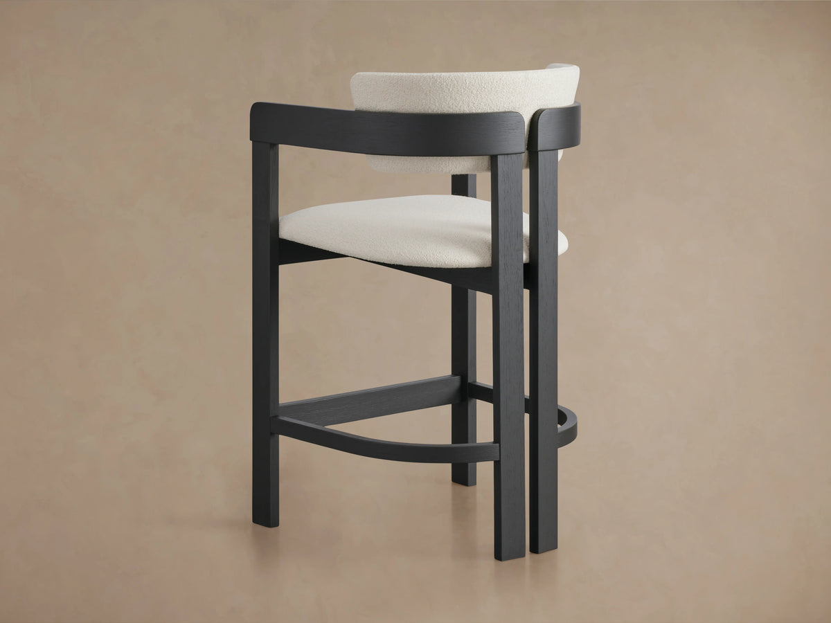 Rodin Counter Stool in a dark ash wood frame with a curved backrest and seat upholstered in light fabric, shown on a beige background.