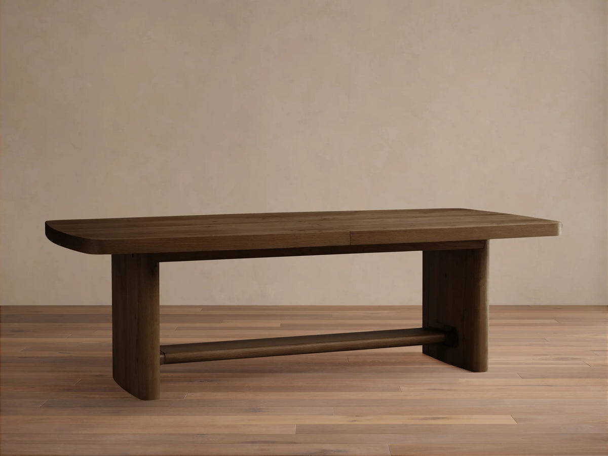 Front view of a brown wooden dining table with an extension leaf seam, rounded corners, and two solid block legs.