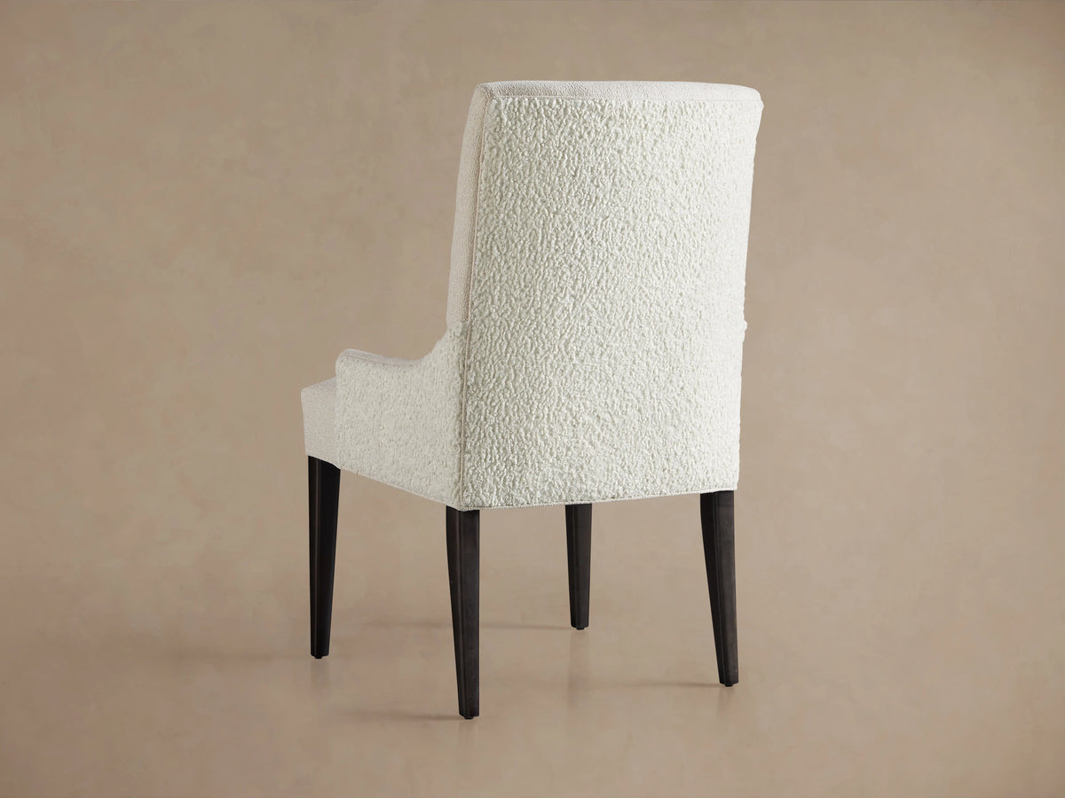 Rhen Dining Chair in soft, textured white fabric with cushioned seat and backrest. Dark wooden legs, set against a beige background.