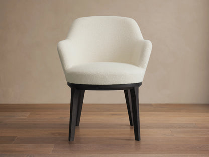 Simone Dining Arm Chair in cream upholstery with a gently curved backrest and armrests, featuring dark wooden legs on a hardwood floor.