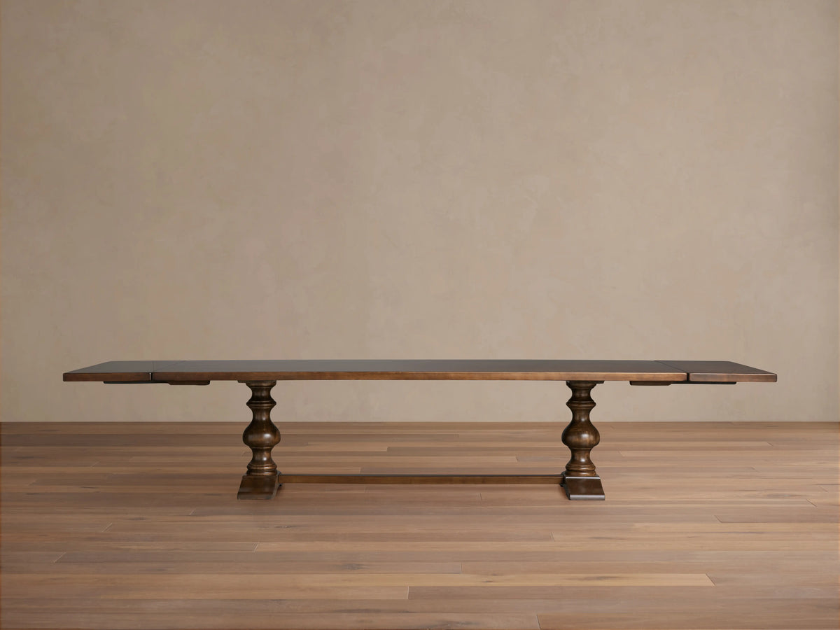 Tuscany Extension Dining Table with a rich, dark wood tabletop, turned pedestal legs, and a horizontal trestle on a wood floor background.