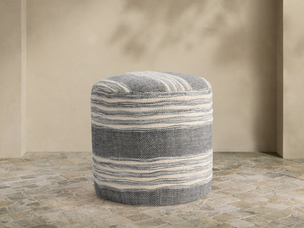 Round performance pouf with horizontal gray and cream stripes on a stone tile floor against a beige wall.