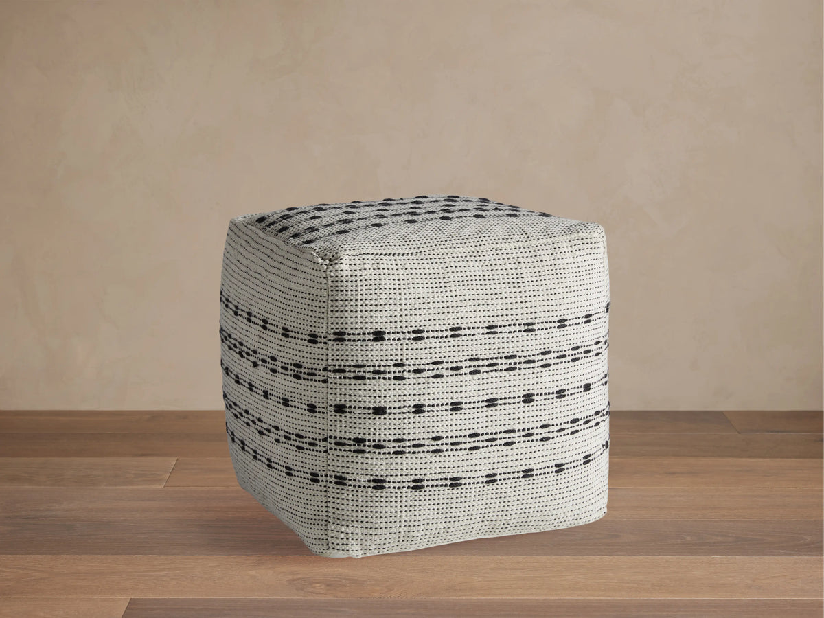 Ladera Performance Pouf with a square shape, featuring a grey and black striped pattern, on a wooden floor against a neutral wall.