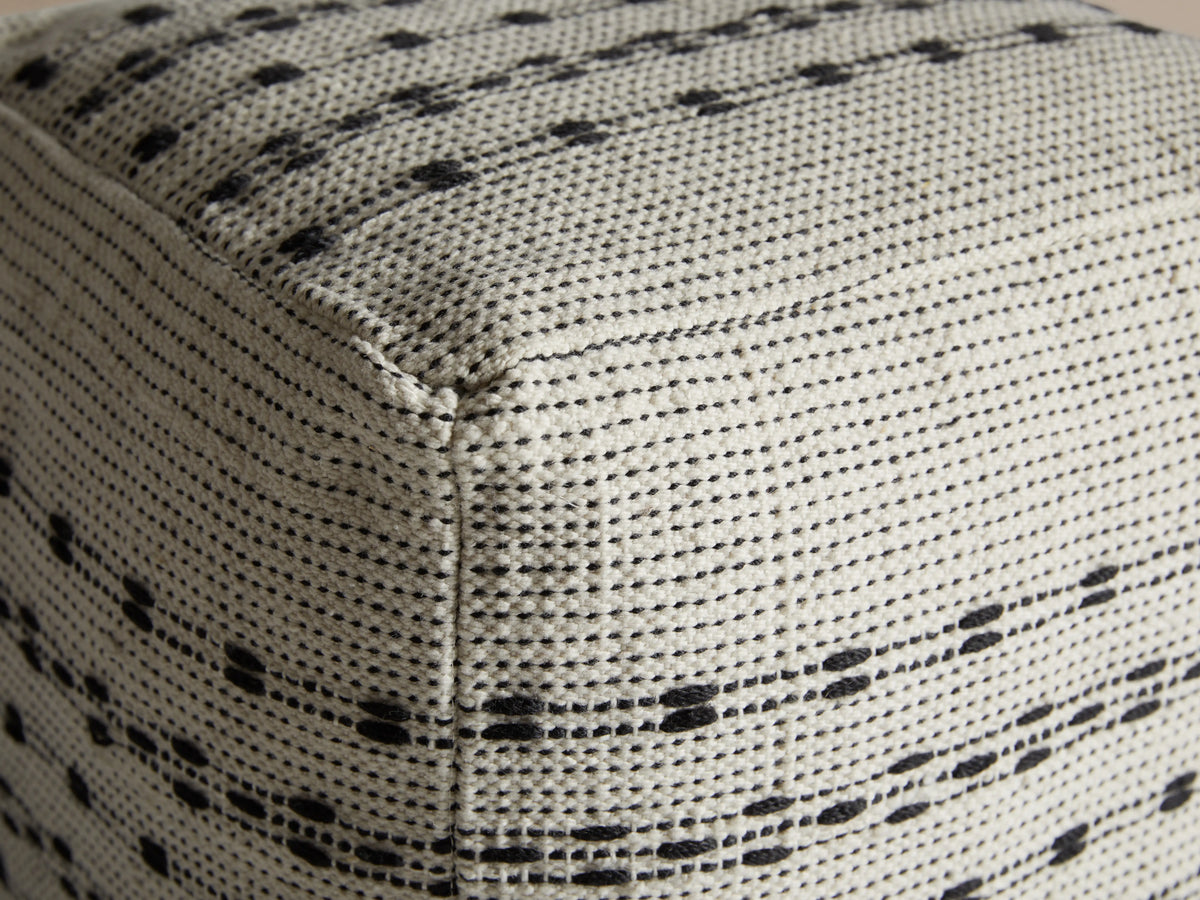 Ladera Performance Pouf with a square shape, featuring a grey and black striped pattern, on a wooden floor against a neutral wall.