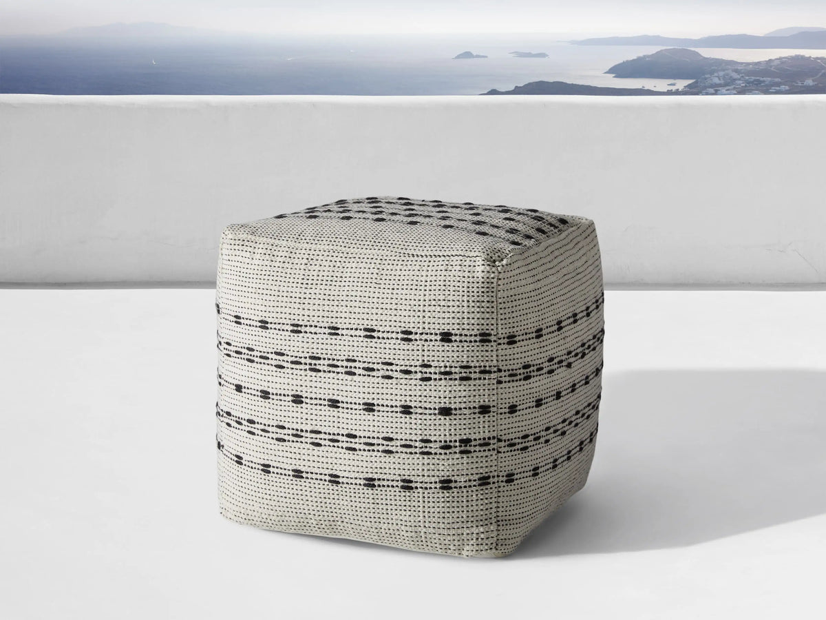 Ladera Performance Pouf, gray with black dotted and striped patterns, cubic shape, placed on wooden floor against beige background.