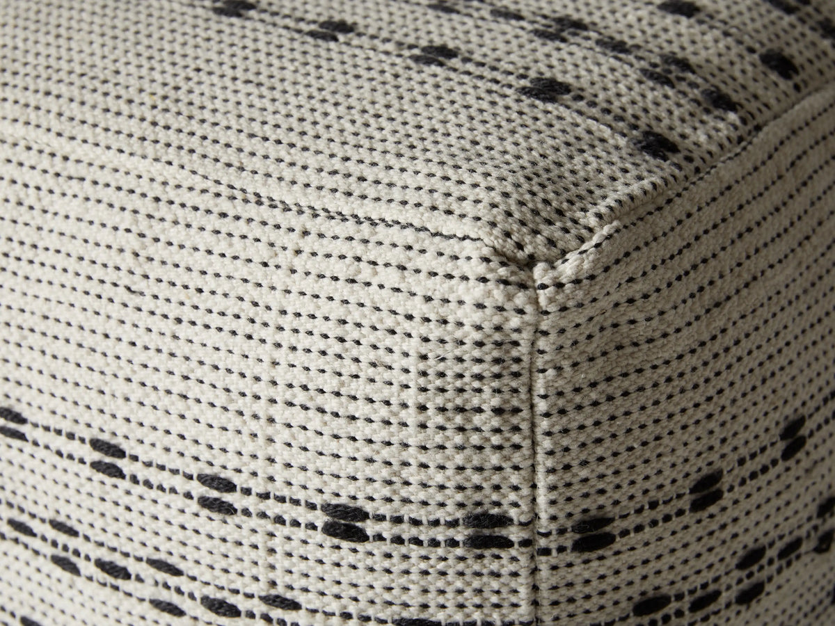 Ladera Performance Pouf in white and black, square-shaped with woven texture and horizontal stripe pattern, placed on a light surface. Ocean view background.