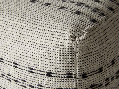 Ladera Performance Pouf in white and black, square-shaped with woven texture and horizontal stripe pattern, placed on a light surface. Ocean view background.