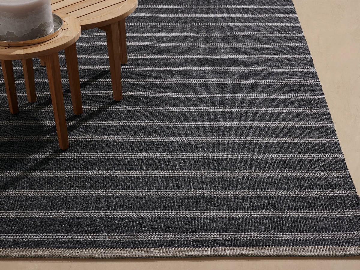 Grover Performance Rug: woven charcoal with thin white horizontal stripes, angled view on a light beige floor, partially under wood tables.