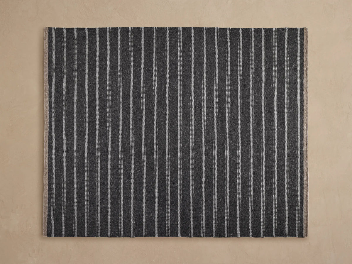 Grover Performance Rug: woven charcoal with thin white horizontal stripes, angled view on a light beige floor, partially under wood tables.