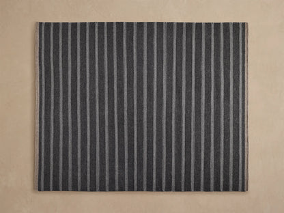 Grover Performance Rug: woven charcoal with thin white horizontal stripes, angled view on a light beige floor, partially under wood tables.