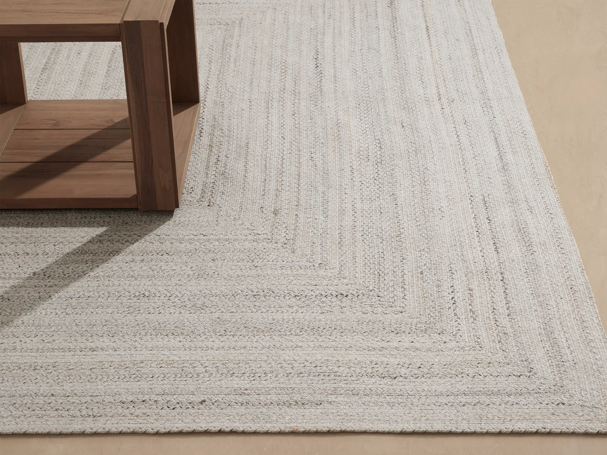 Sansa Performance Rug: light grey braided rug with a rectangular border, partially visible under a wooden table.