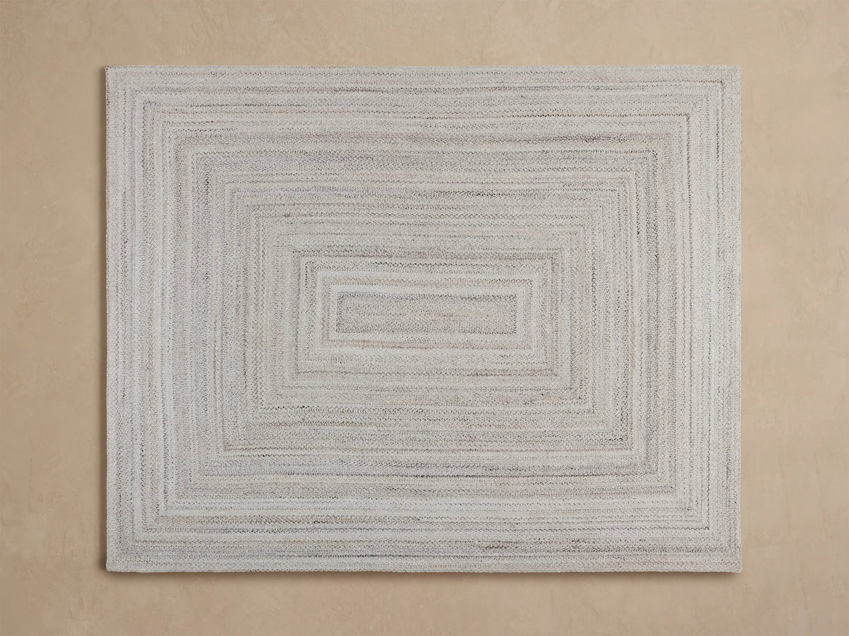Sansa Performance Rug: light grey braided rug with a rectangular border, partially visible under a wooden table.