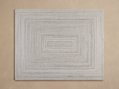 Sansa Performance Rug: light grey braided rug with a rectangular border, partially visible under a wooden table.