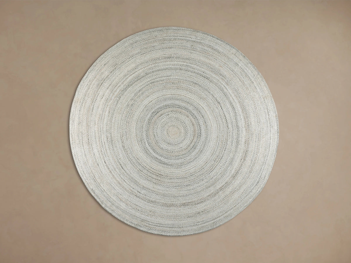 Sansa Performance Round Rug, a light gray braided round rug with concentric circles, on a wooden floor beside furniture.