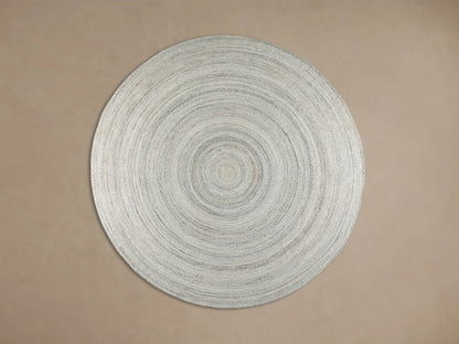 Sansa Performance Round Rug, a light gray braided round rug with concentric circles, on a wooden floor beside furniture.