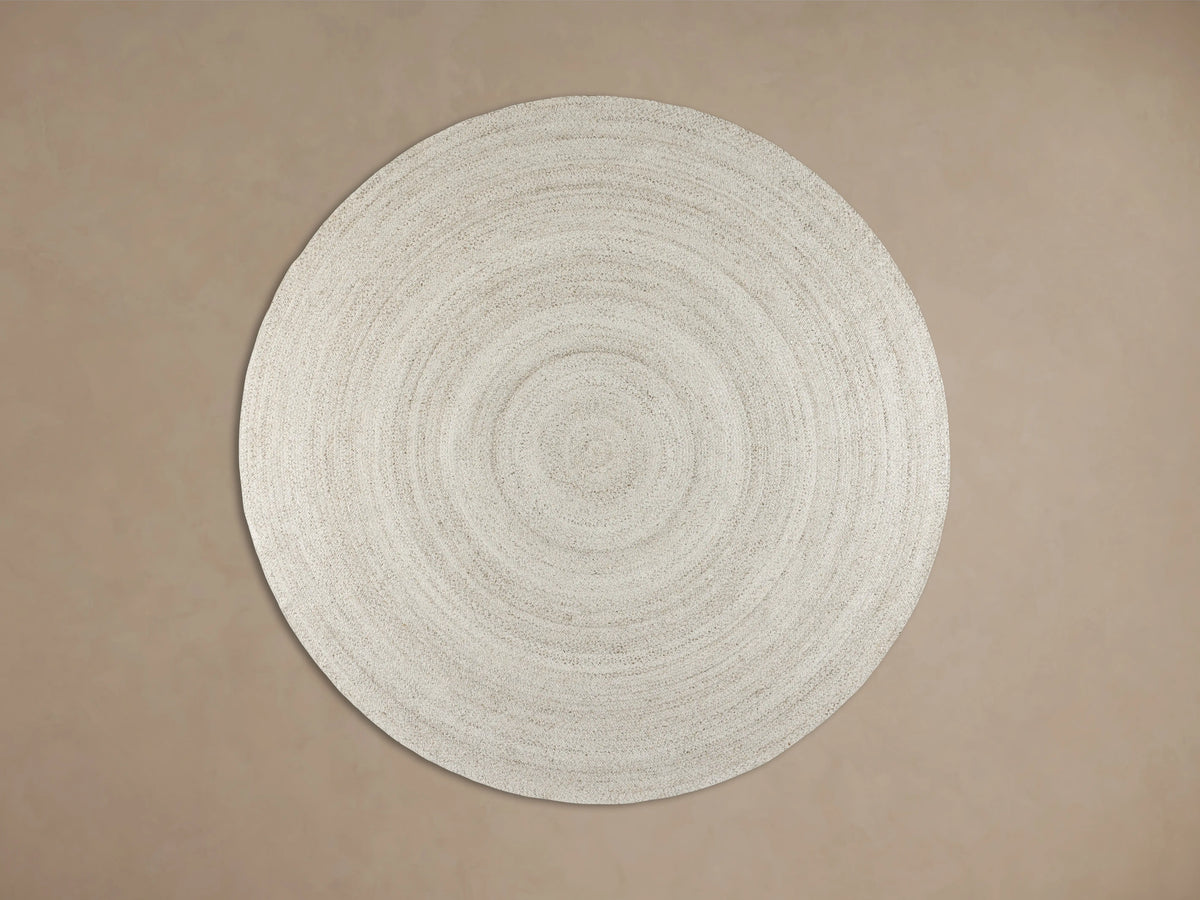 Sansa Performance Round Rug, natural braided with concentric circles, partly under black furniture on wood floor.