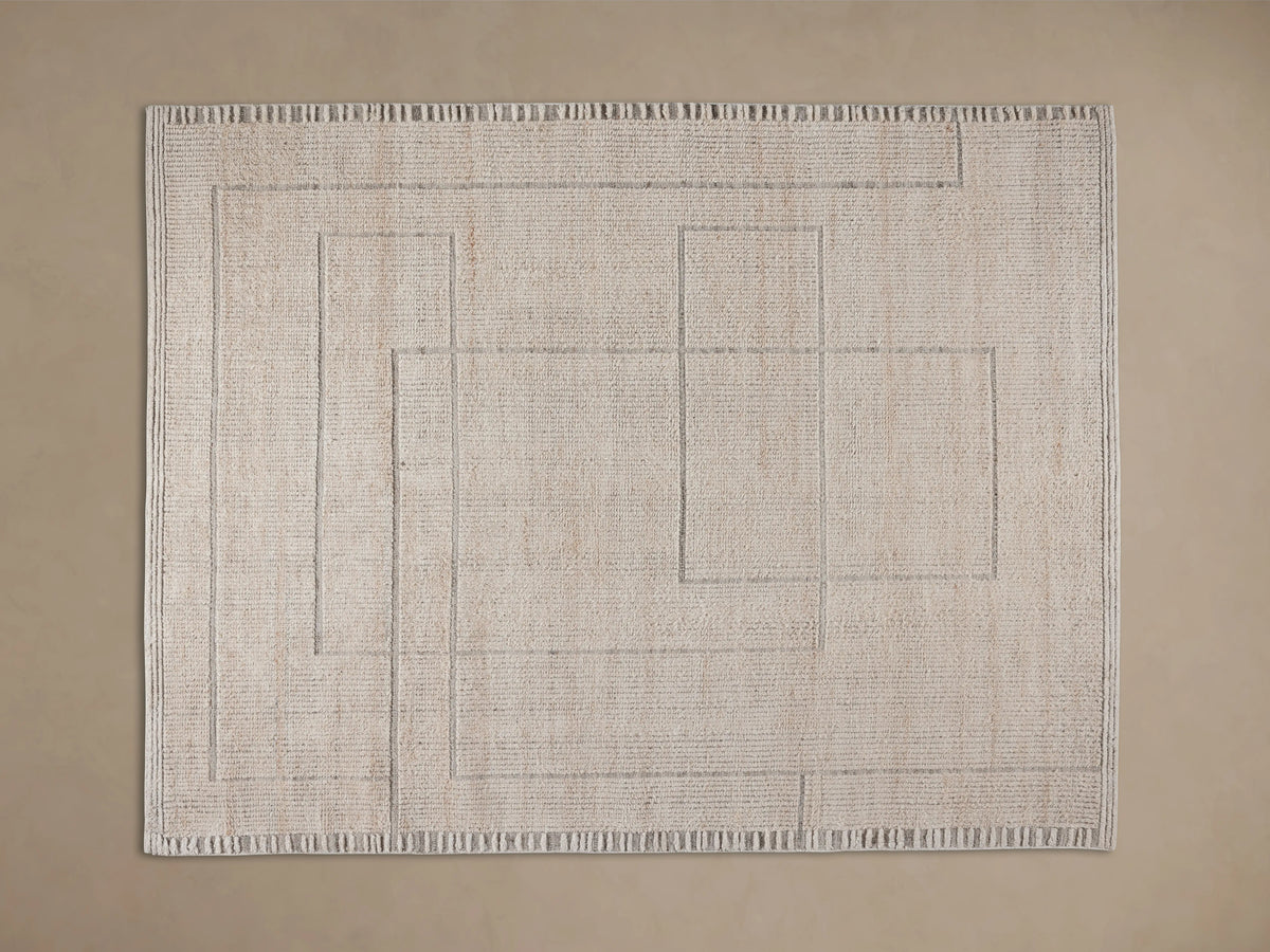 Close-up of a light beige Walker Performance Rug with raised linear and geometric patterns, partially under a wooden table on a wood floor.