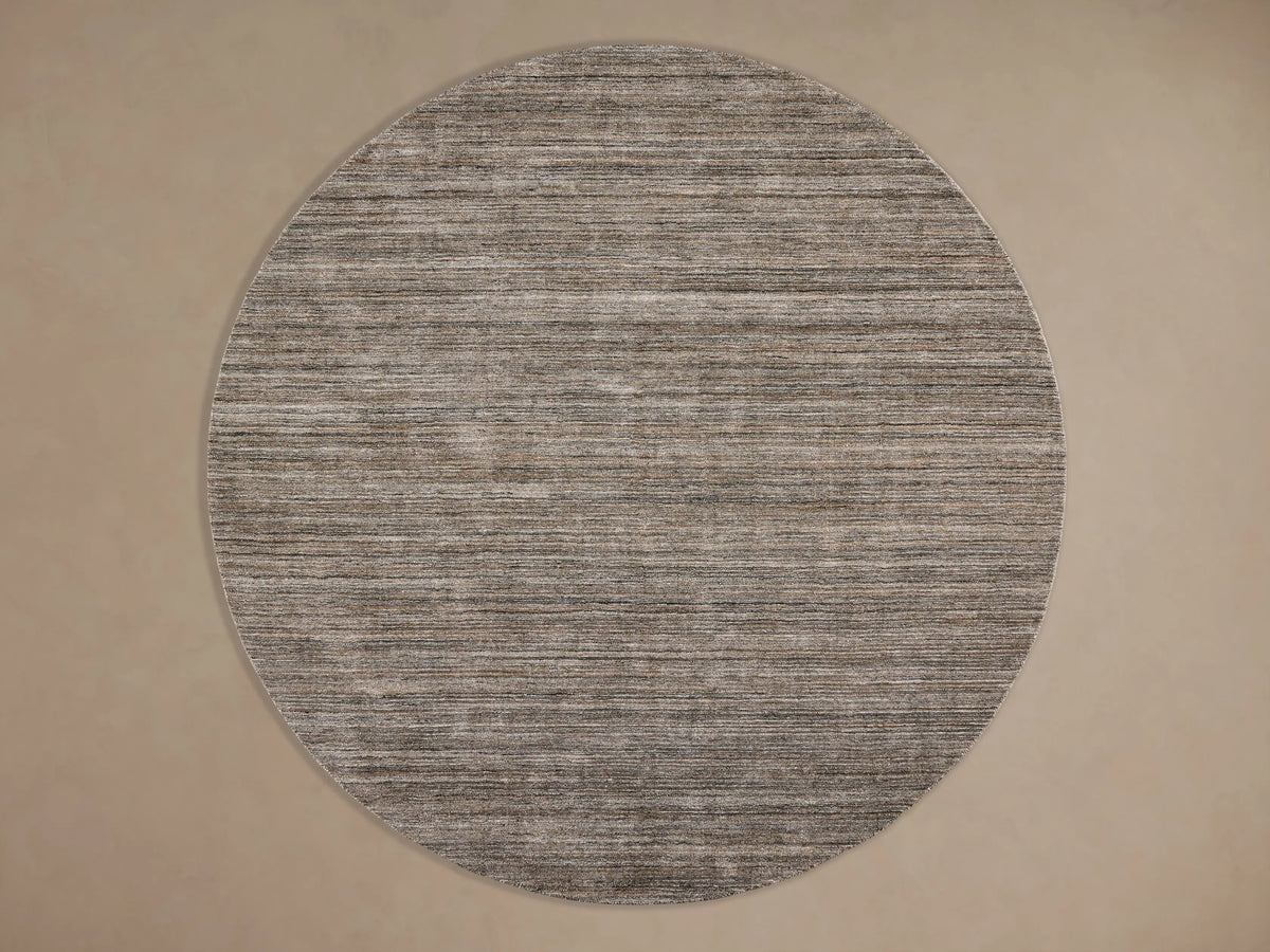 Textured round rug with horizontal stripes in multi-tone gray and brown on a dark wood floor, with a dark coffee table partially visible.