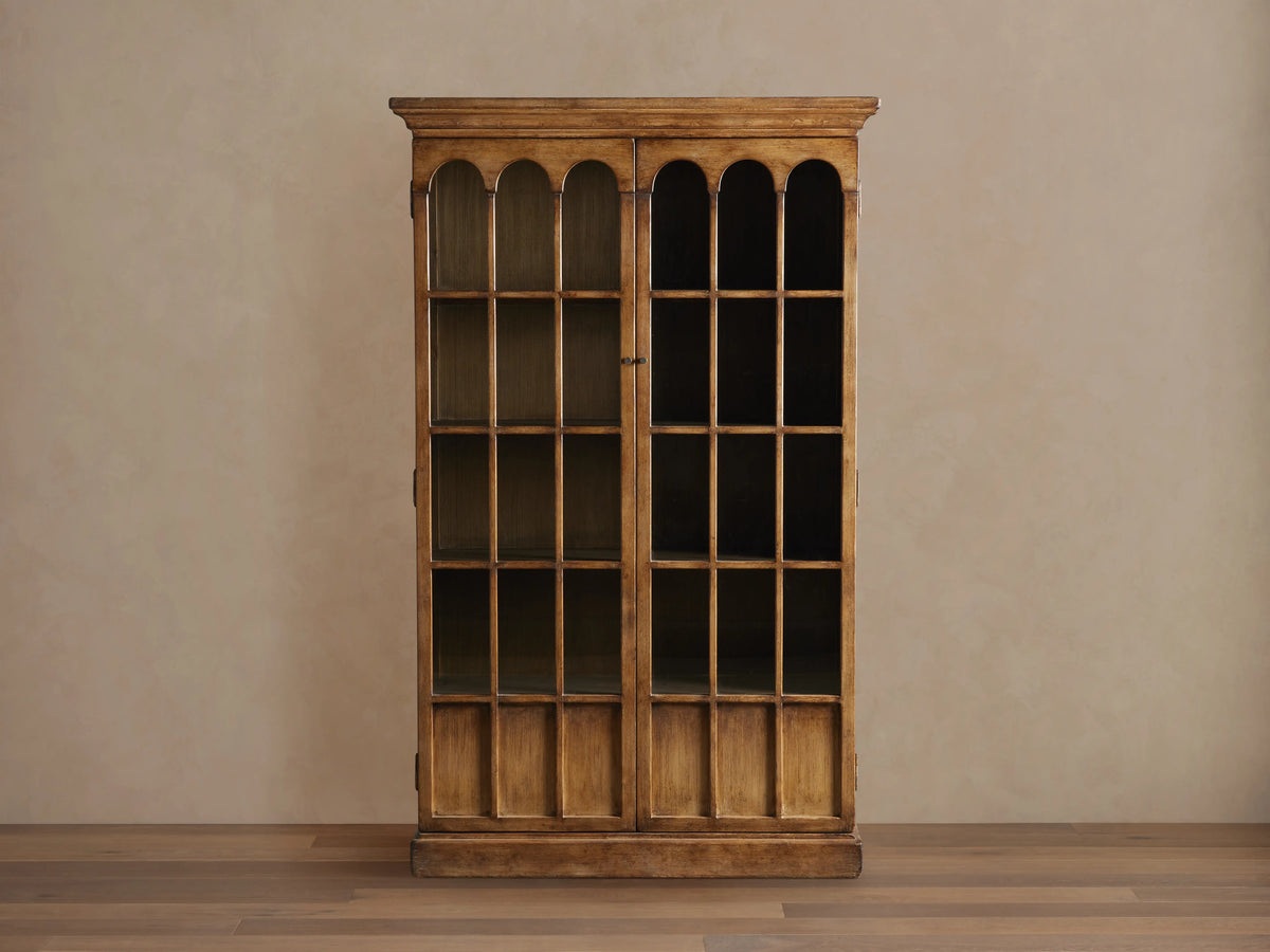 Merritt Cabinet in distressed pine with two glass doors featuring arched panes, antiqued brass hardware, and a warm lacquer finish.