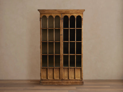 Merritt Cabinet in distressed pine with two glass doors featuring arched panes, antiqued brass hardware, and a warm lacquer finish.
