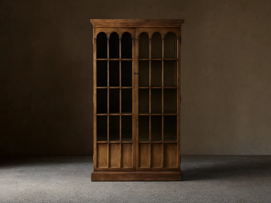 Merritt Cabinet in distressed pine with two glass doors featuring arched panes, antiqued brass hardware, and a warm lacquer finish.