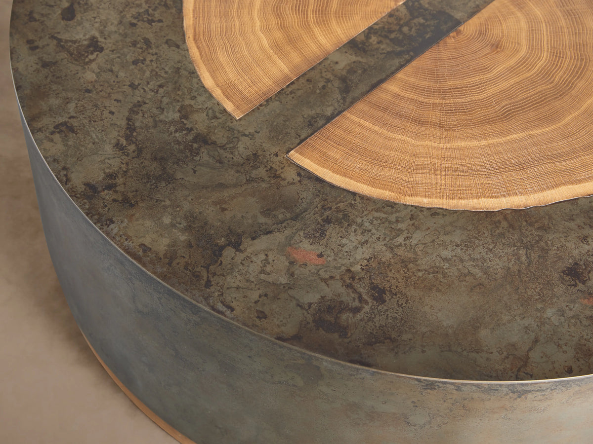 Round coffee table with a dark oxidized steel body and light wood accents. Top features concentric veneer circles from reclaimed wood.
