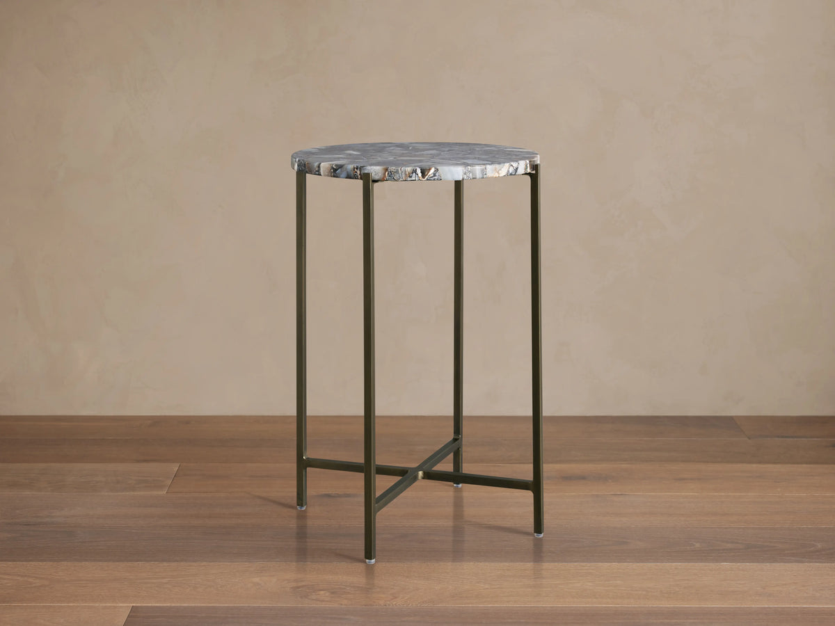Ludlow Martini Table with a round, polished stone top and slender, hand-welded iron legs, on a wooden floor background.