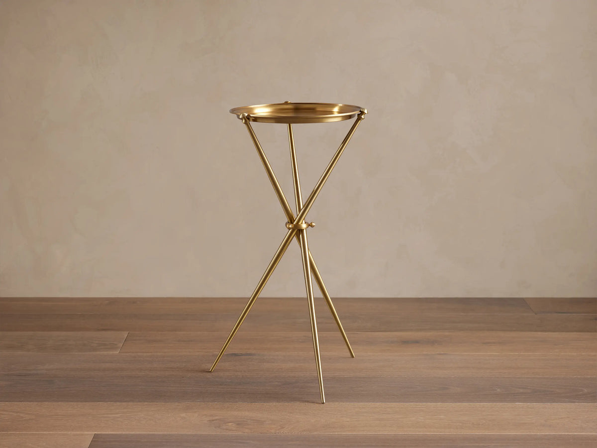 Albie Martini Table with a sleek gold finish, features a round top and a tripod base, set on a wooden floor against a beige wall.