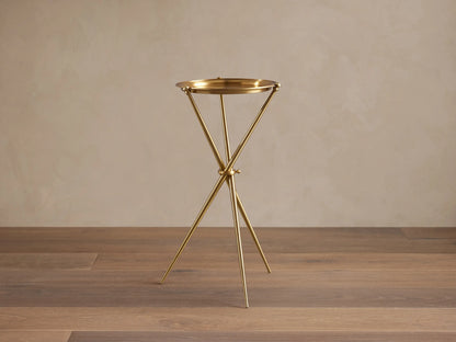Albie Martini Table with a sleek gold finish, features a round top and a tripod base, set on a wooden floor against a beige wall.
