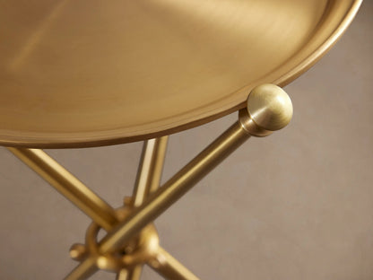 Albie Martini Table with a sleek gold finish, features a round top and a tripod base, set on a wooden floor against a beige wall.